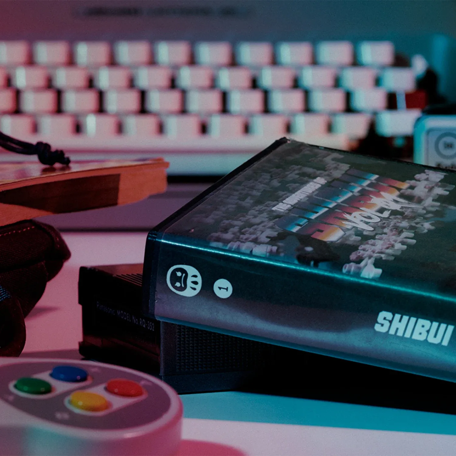 Videocassetta Shibui, tastiera e controller su una scrivania illuminata di rosa e azzurro